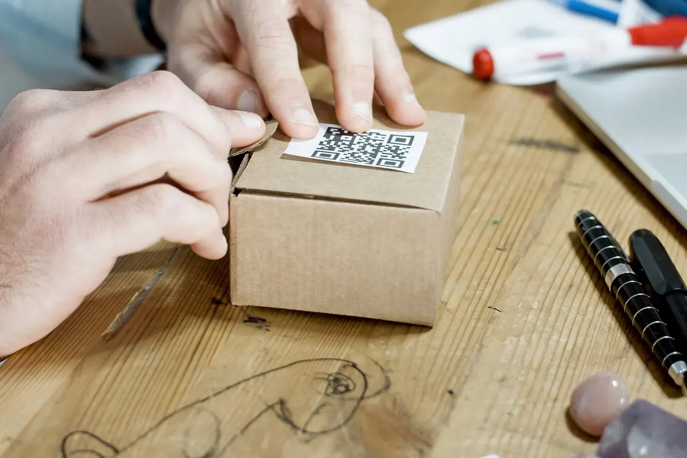 Hands opening a small cardboard parcel with a label on its lid, on a wooden table.